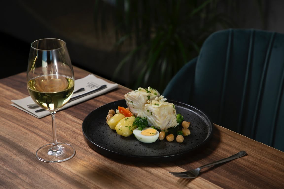 Bacalhau cozido com grão, ovo e batata num prato preto, acompanhado de copo de vinho branco