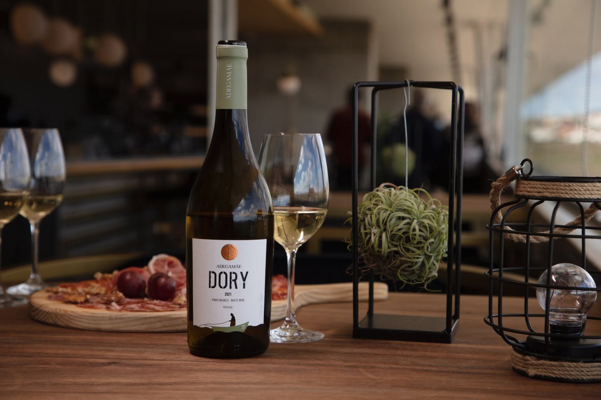 Garrafa de vinho branco AdegaMãe Dory 2021 com copo e tábua de charcutaria