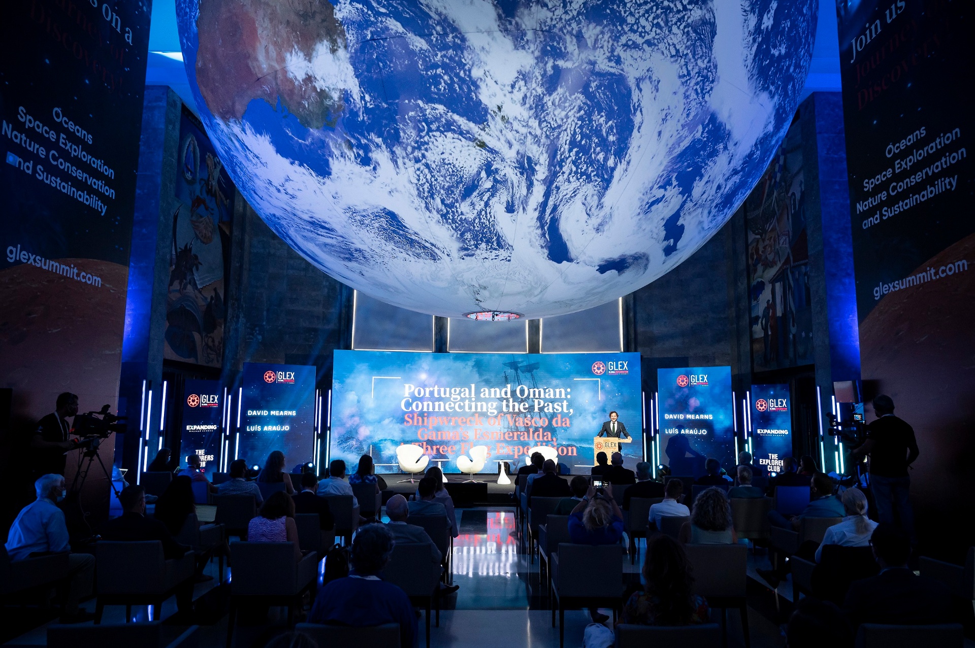 Sessão da GLEX Summit sob um globo terrestre suspenso, com plateia e palco iluminados a azul