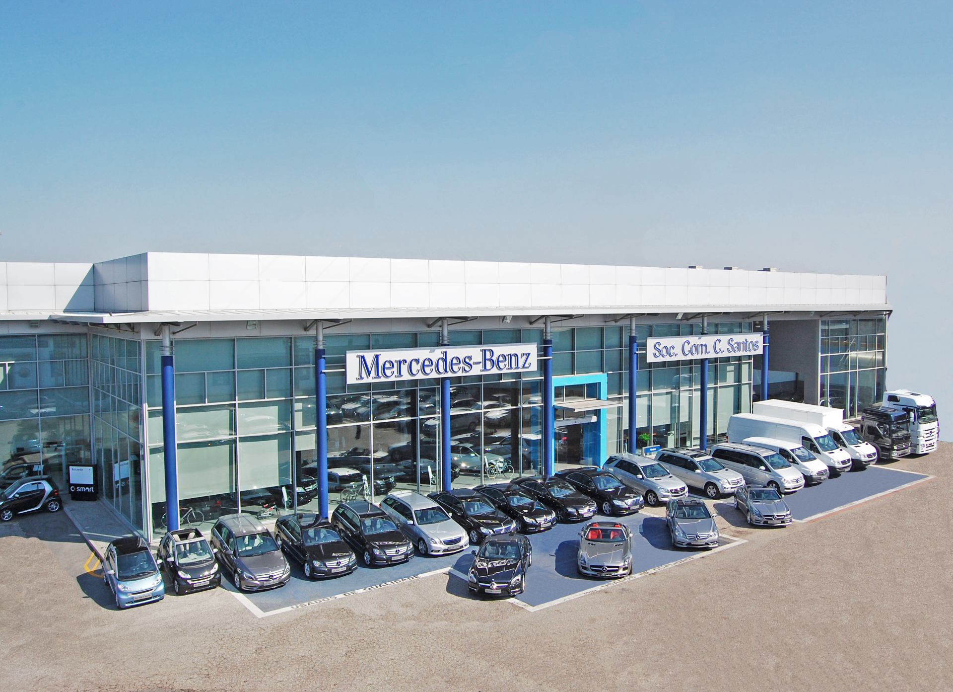 Vista exterior das instalações da Sociedade Comercial C. Santos, concessionário Mercedes-Benz e smart, com fachada envidraçada, pilares azuis e gama de viaturas exposta na frente