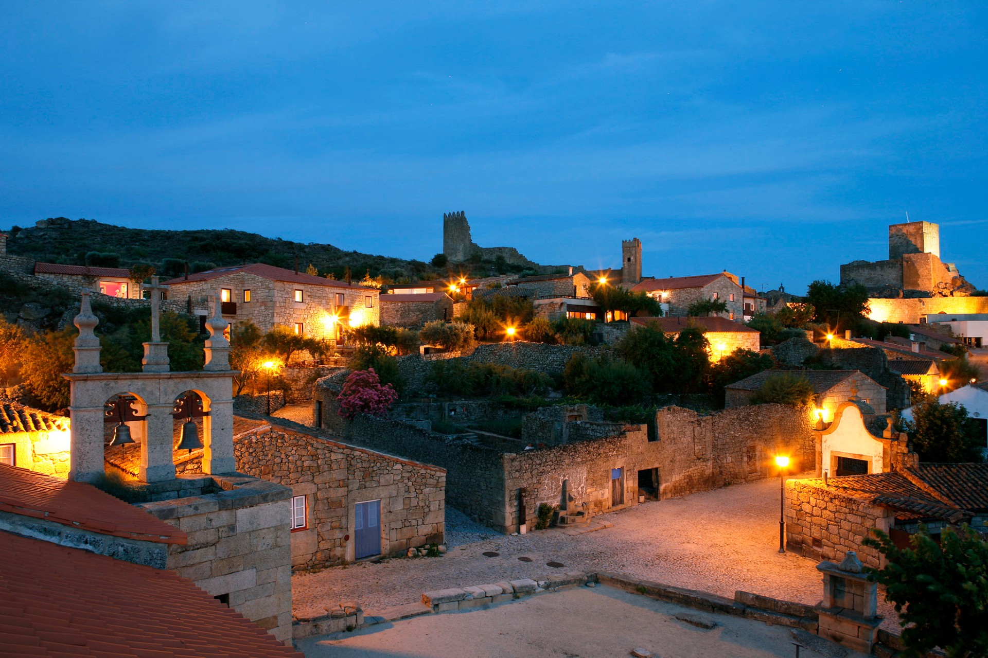 Vista panorâmica de aldeia histórica ao anoitecer com castelo iluminado ao fundo e candeeiros acesos entre casas de granito