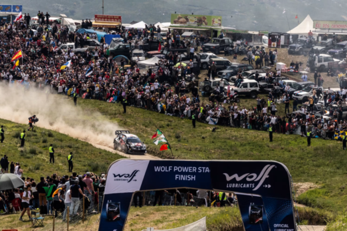 Vista panorâmica da Wolf Power Stage com público, viaturas estacionadas e um carro de rally a cortar a meta envolto em poeira