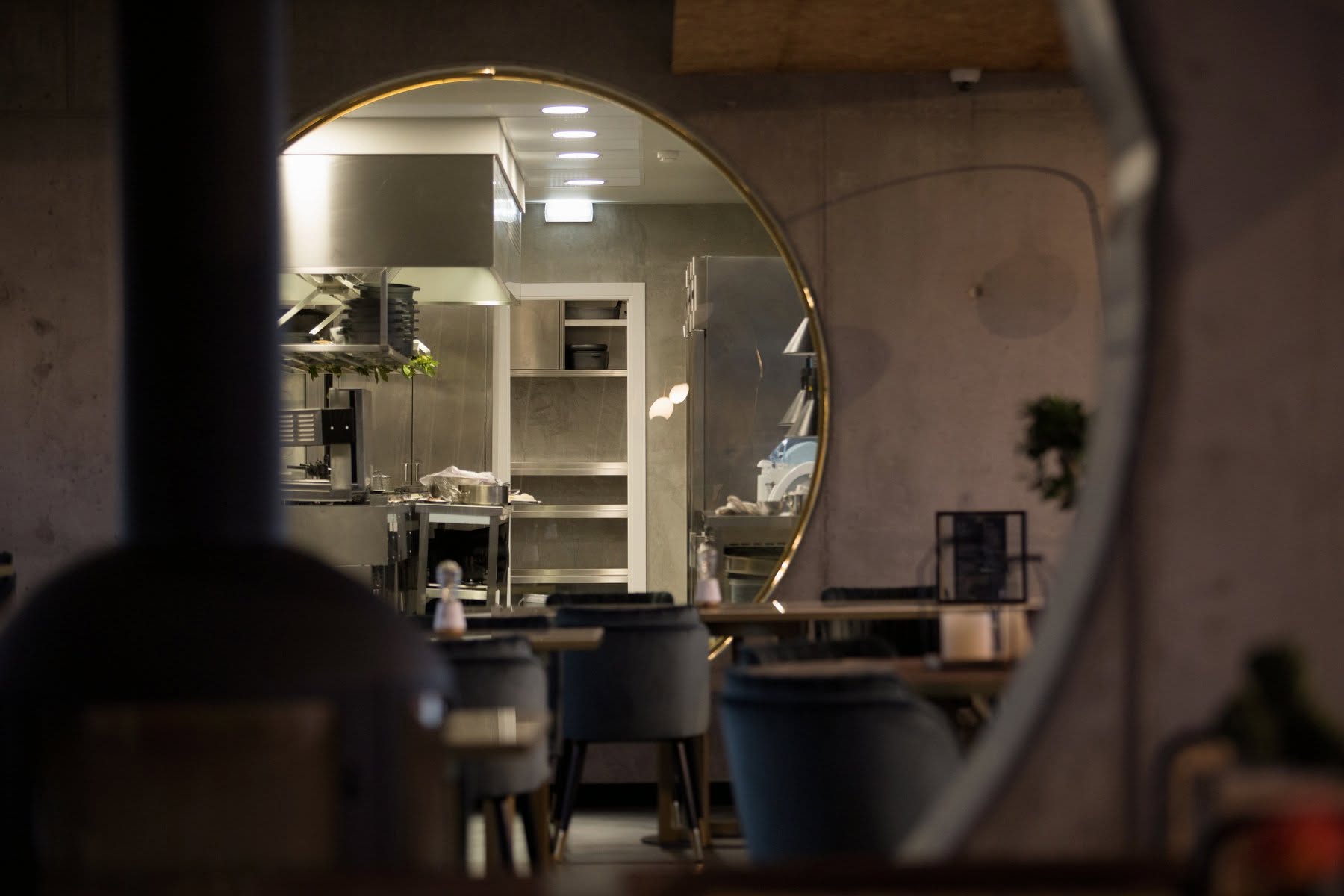 Interior do restaurante Sal na Adega visto através de espelho redondo dourado, com cozinha aberta ao fundo