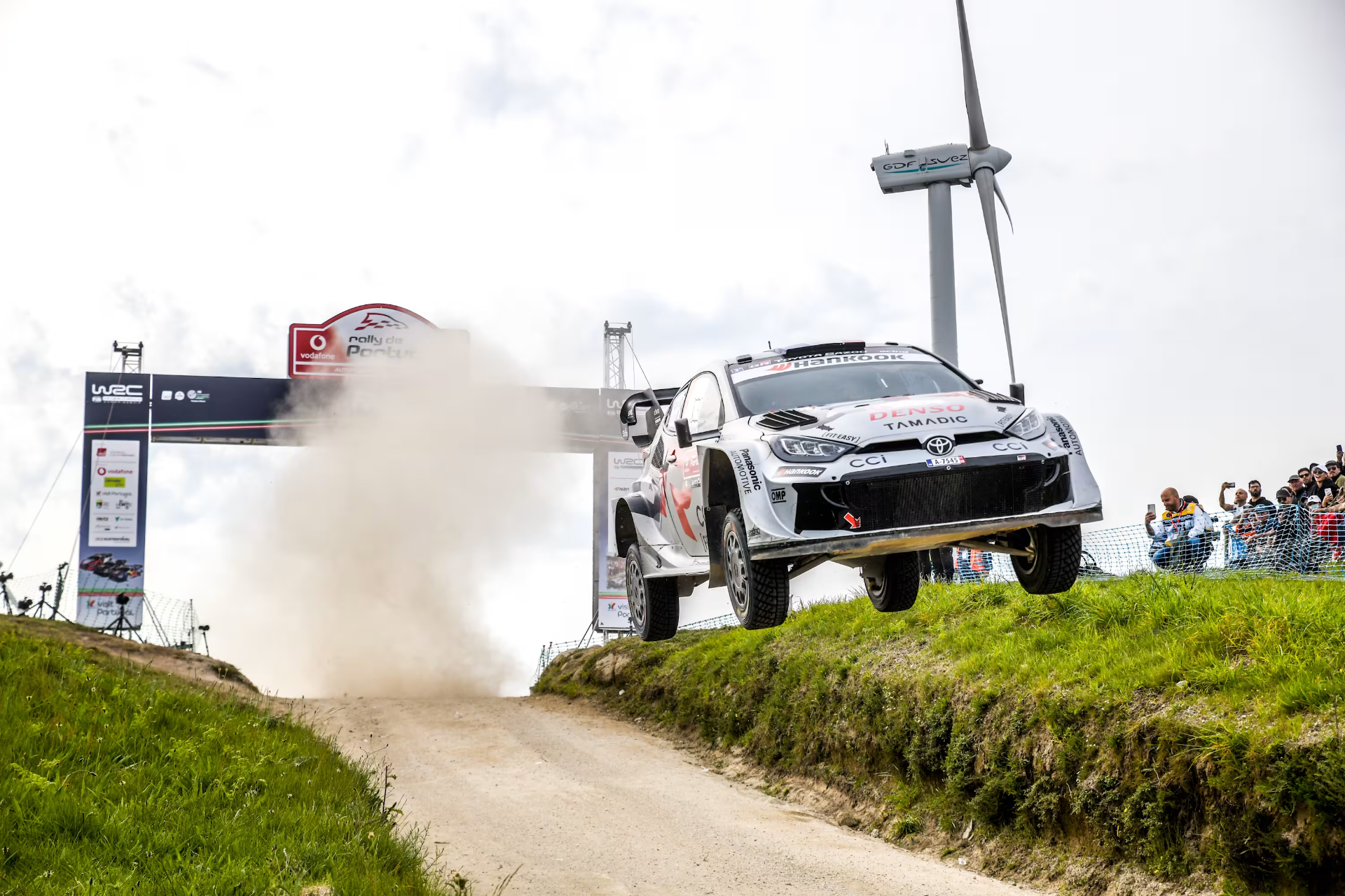 Toyota GR Yaris em pleno salto sobre uma classificativa de terra com aerogerador ao fundo