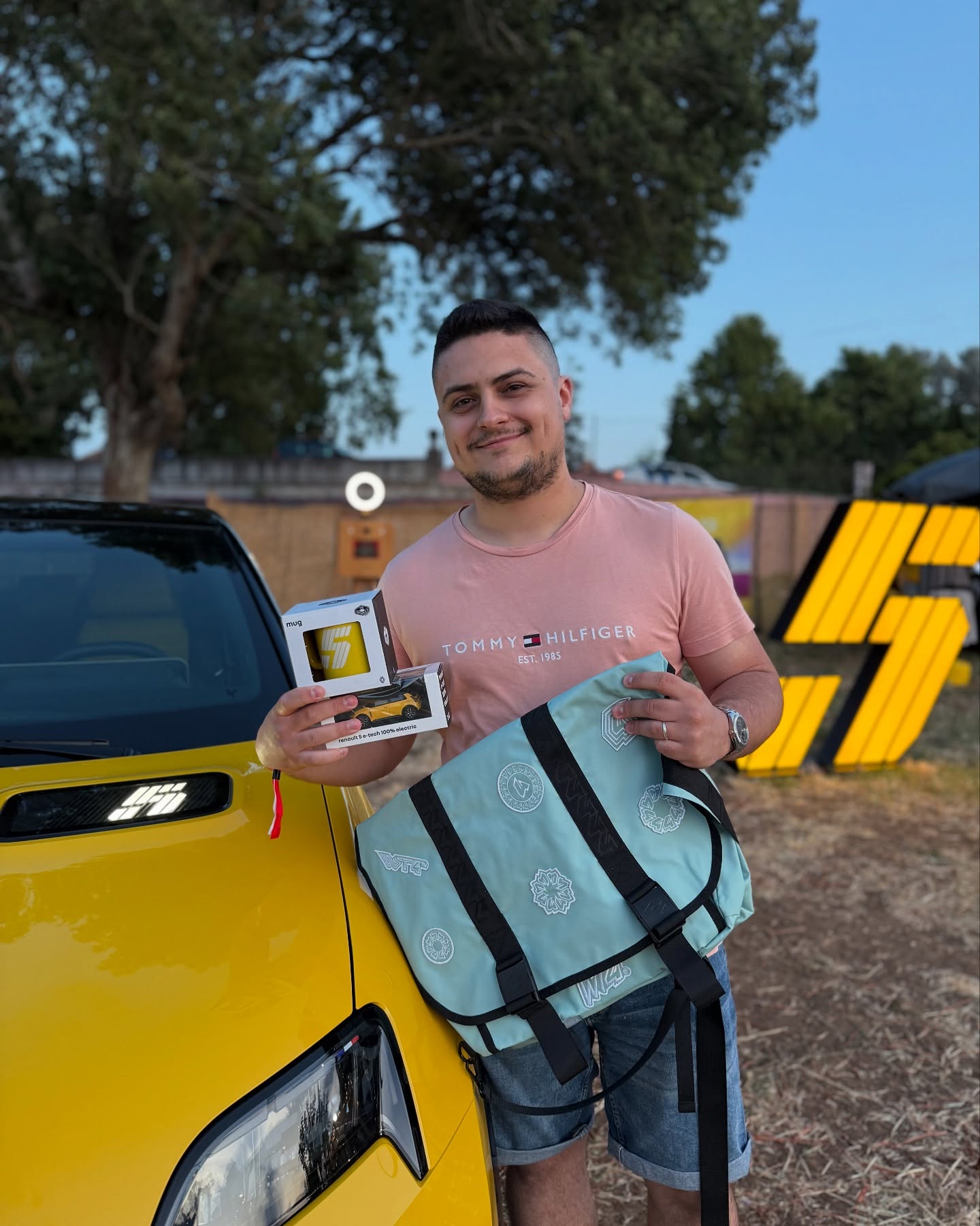 Visitante a posar com brindes Renault 5 — caneca, miniatura e mochila — junto a um Renault 5 amarelo