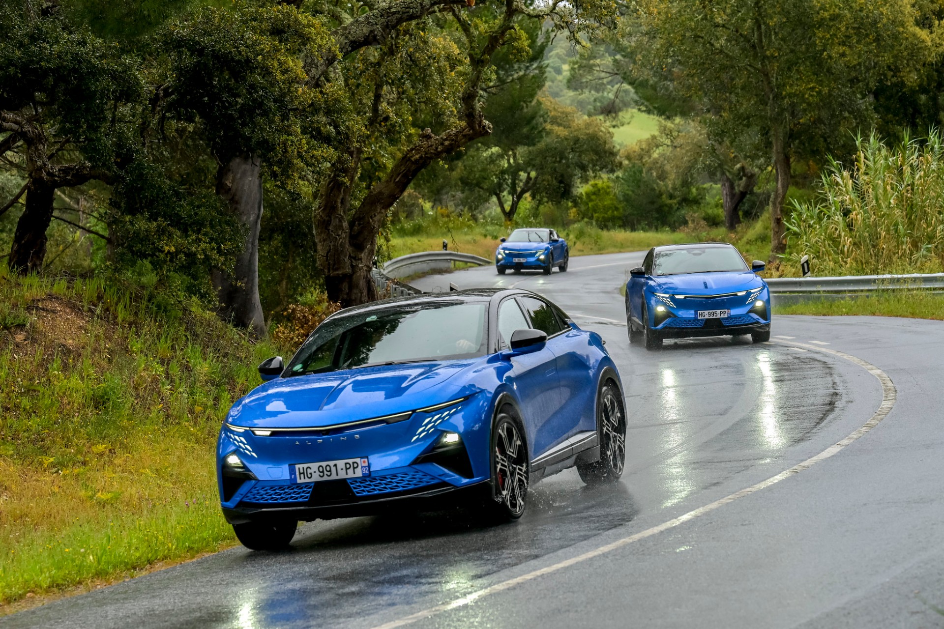 Três Alpine A390 GT azuis em comboio numa curva de estrada nacional molhada, entre sobreiros do Alentejo