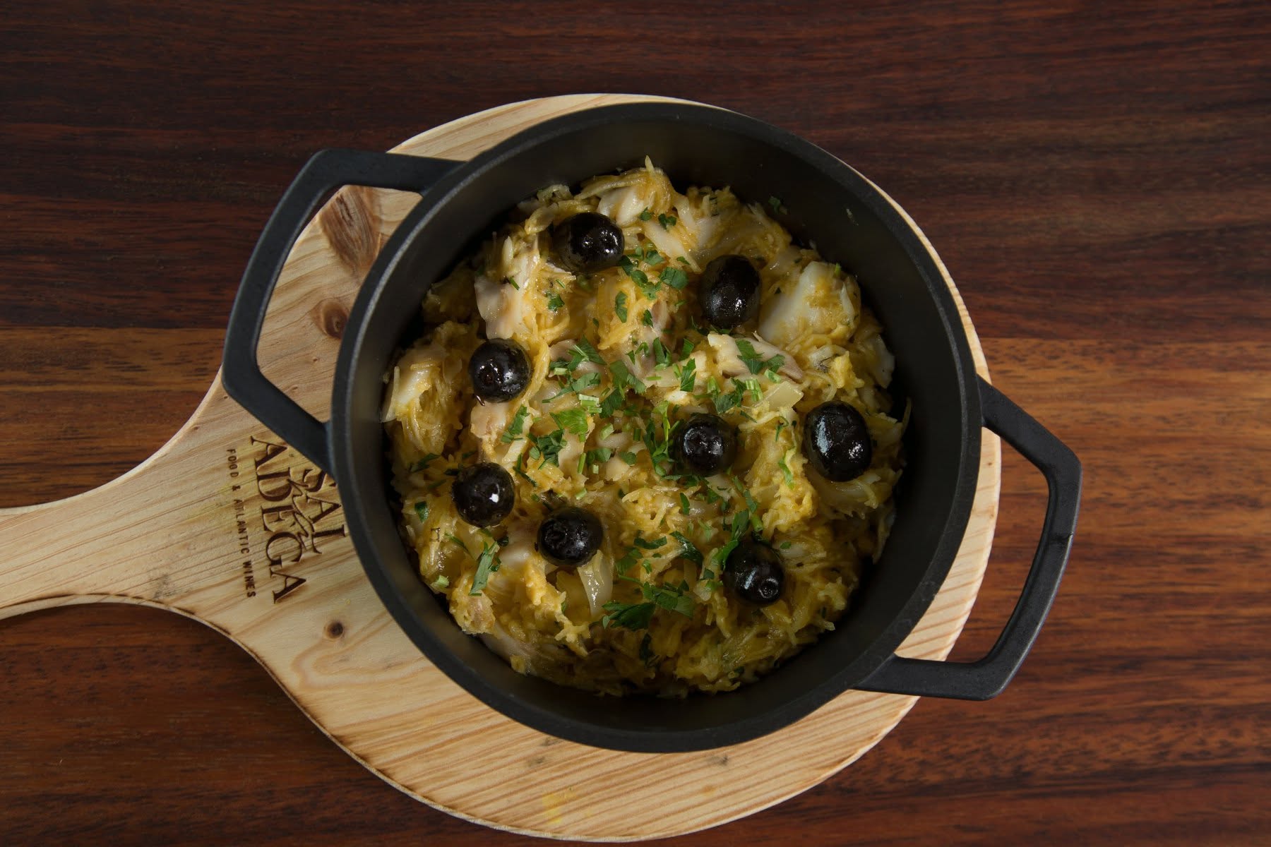 Bacalhau à Brás com azeitonas pretas e salsa em tacho de ferro fundido sobre tábua Sal na Adega