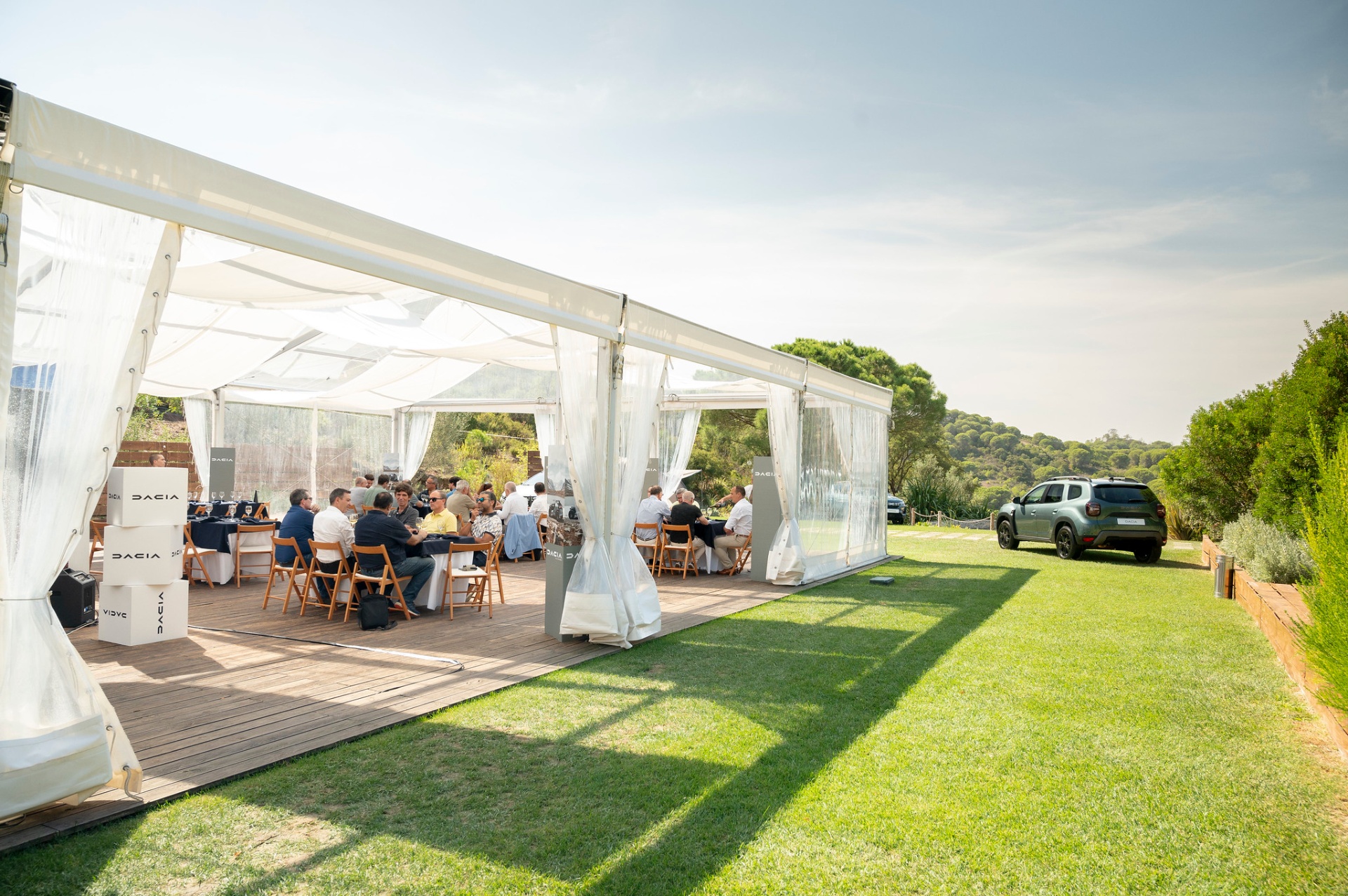 Vista exterior da tenda transparente com mesas de almoço e Dacia Duster ao fundo