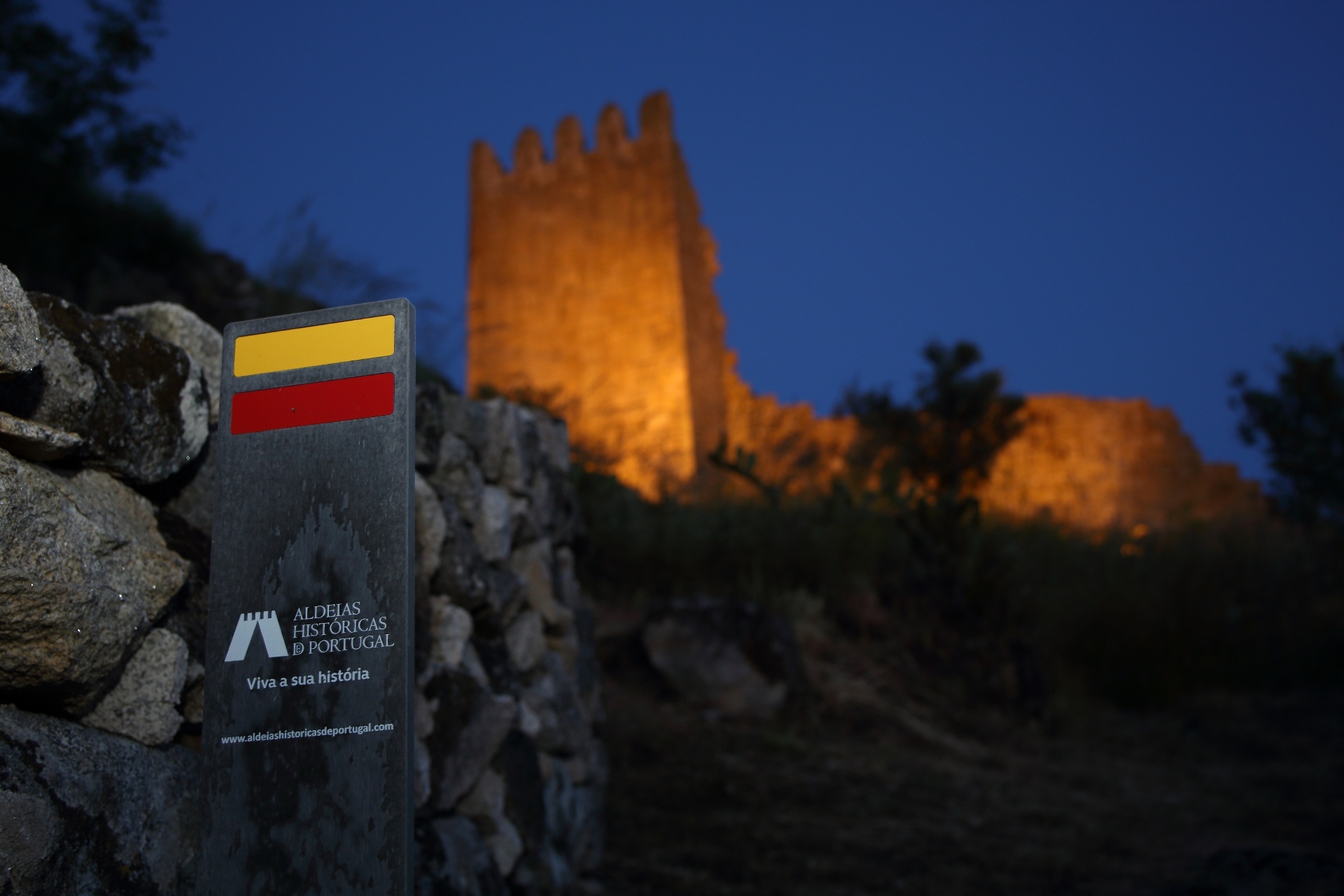 Sinalética das Aldeias Históricas de Portugal junto a muro de pedra com torre de castelo iluminada ao anoitecer