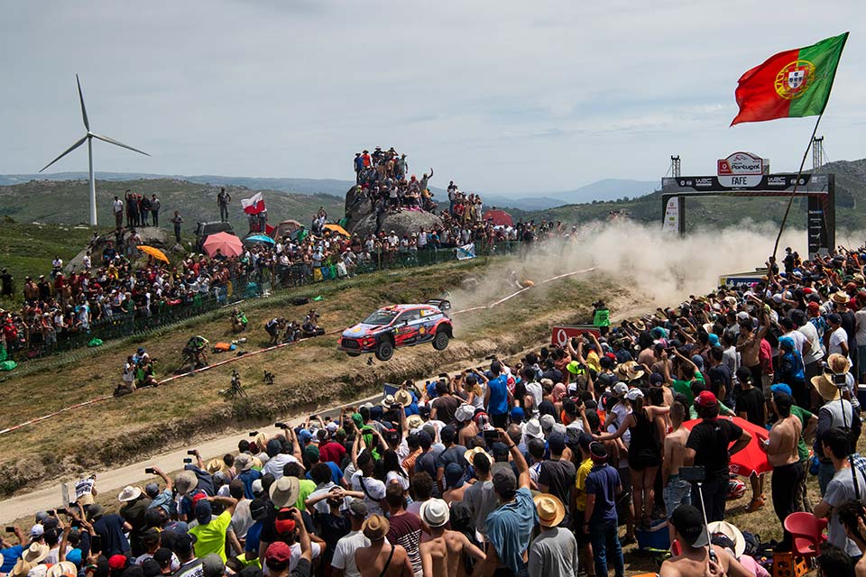 Multidão no salto de Fafe com bandeira de Portugal em destaque e um WRC a passar a alta velocidade levantando poeira