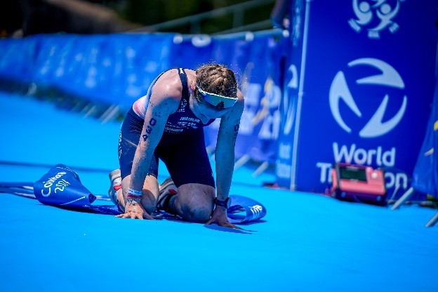 Atleta exausta de joelhos na zona azul de chegada com lona World Triathlon ao fundo