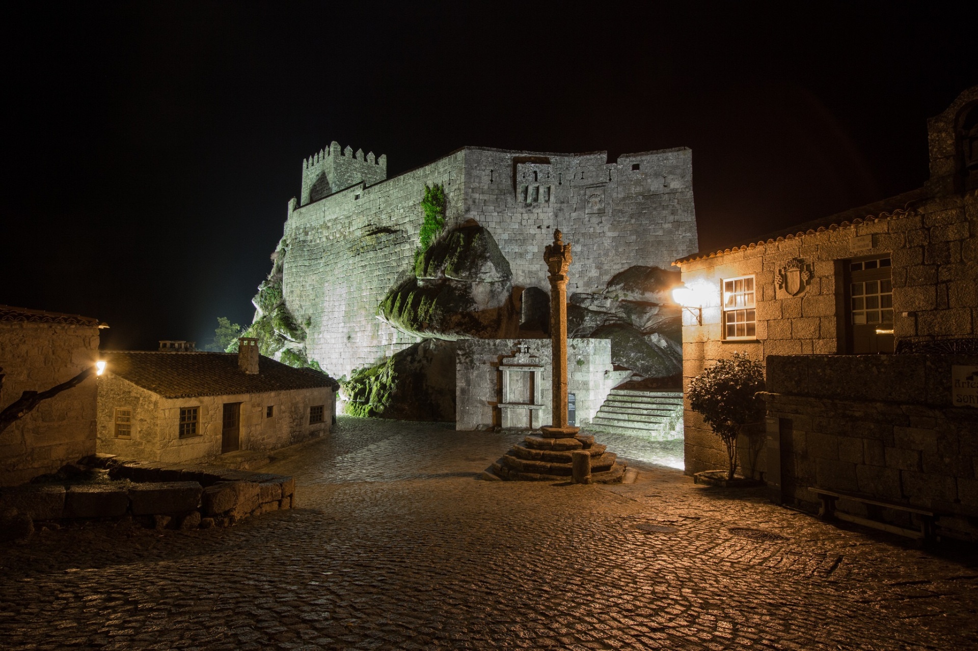 Praça de Sortelha à noite com pelourinho em primeiro plano e castelo iluminado sobre rochedos ao fundo